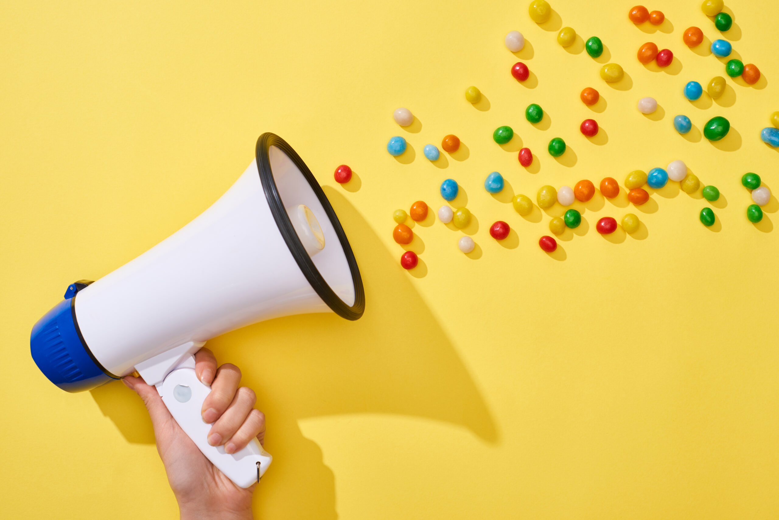 a woman holds a megaphone on a yellow background; the megaphone has rainbow candy coming out of it