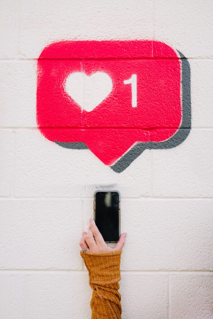 A woman holds a phone up to a white wall where 1 like is drawn on in chalk
