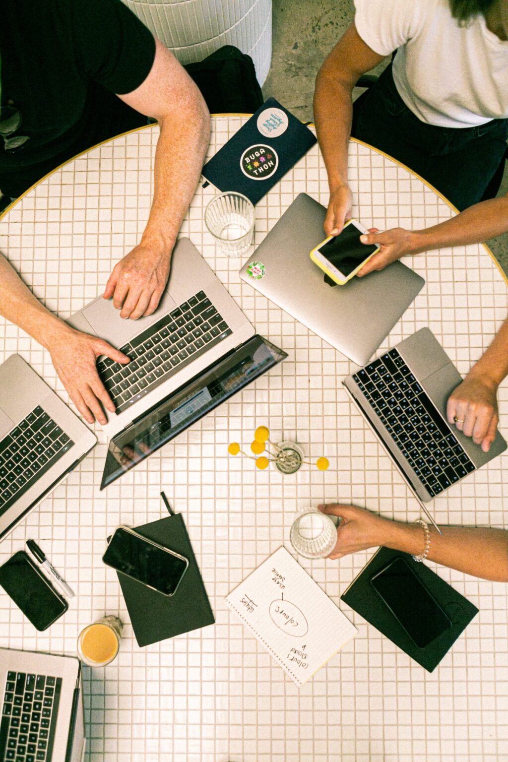 A desk with four laptops, three notebooks, iPhones, and people's arms on a desk are shown from a bird's eye view.